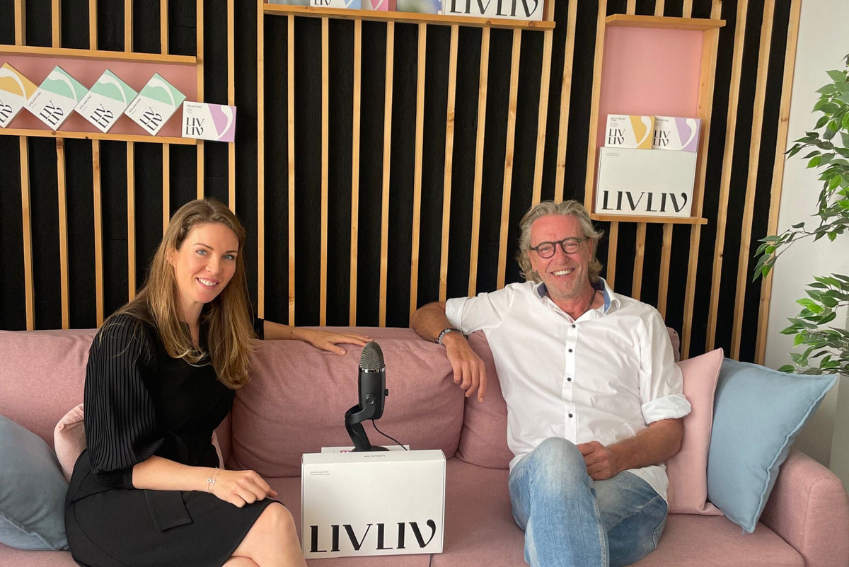 Man en vrouw zitten samen op een roze bank voor podcast over gezonde levensstijl, microfoon op tafel, doos met LIVLIV producten zichtbaar.